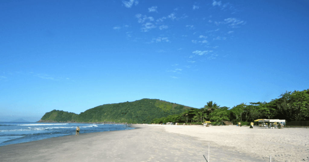 Imagem da Praia de Cambury, em São Sebastião, mostrando a extensa faixa de areia clara, o mar calmo com pequenas ondas, o morro verde ao fundo e quiosques espalhados próximos à vegetação nativa, destacando o clima tranquilo e preservado do litoral norte paulista.