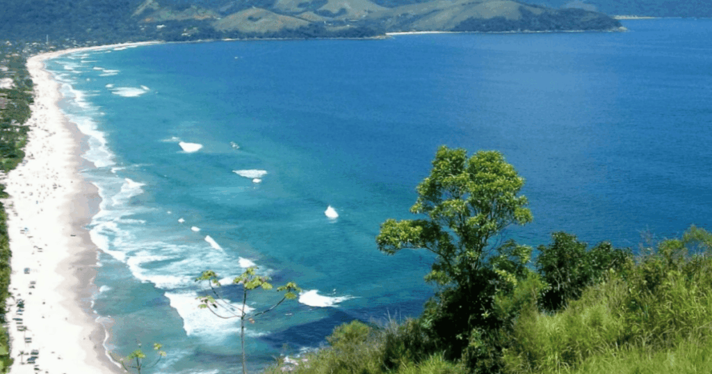 Vista panorâmica da Praia de Maresias, em São Sebastião, mostrando o mar azul intenso, ondas suaves chegando à faixa de areia clara e vegetação verde em primeiro plano, destacando a beleza do litoral norte de São Paulo.
