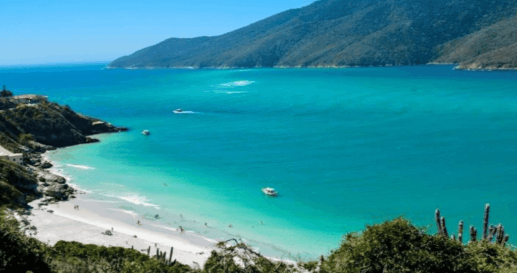 Vista aérea das Prainhas do Pontal do Atalaia, em Arraial do Cabo, com mar azul-turquesa em diferentes tons, areia branca fina, barcos ancorados e morros cobertos por vegetação ao fundo.