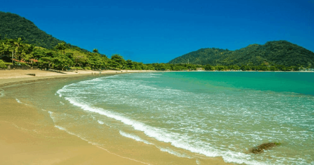 Foto da Praia do Lázaro (Ubatuba), mostrando o mar verde marinho, com pequenas montanhas ao fundo e a extensa faixa de areia, no litoral norte de SP 
