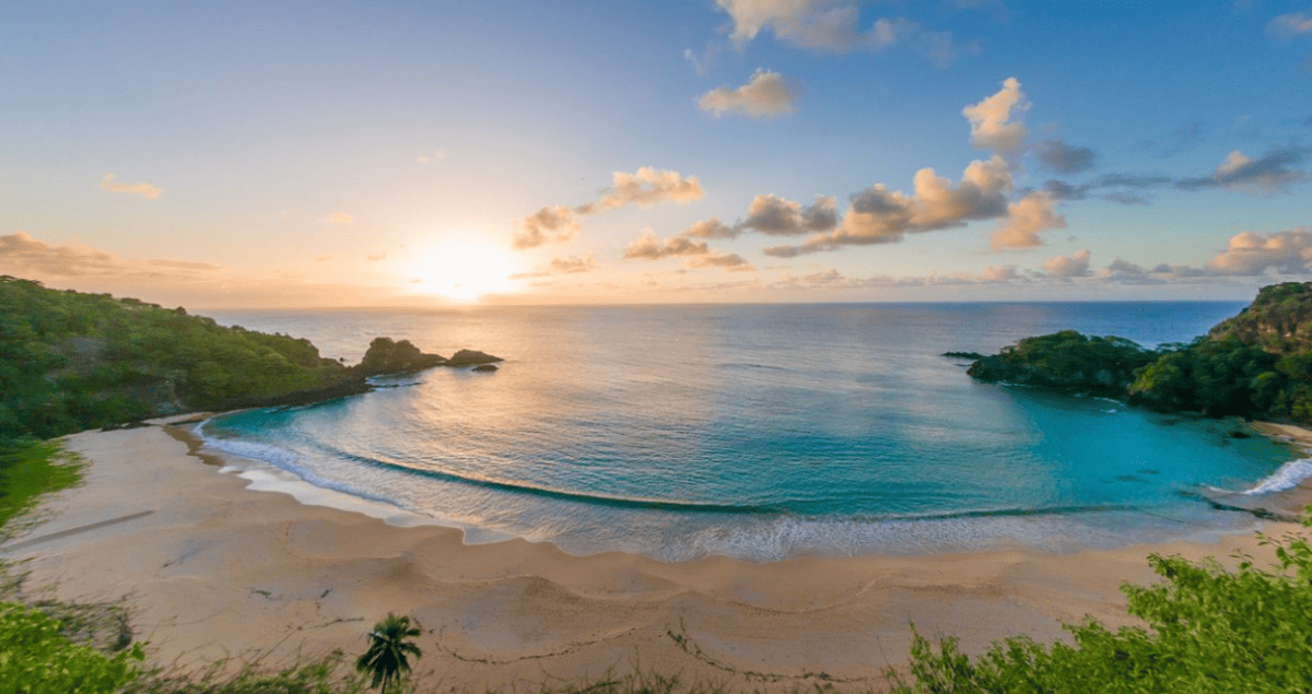 Vista panorâmica da Baía do Sancho em Fernando de Noronha, com mar azul cristalino, falésias cobertas por vegetação e o sol se pondo no horizonte, iluminando a praia de forma suave, umas das praias mais bonitas do brasil