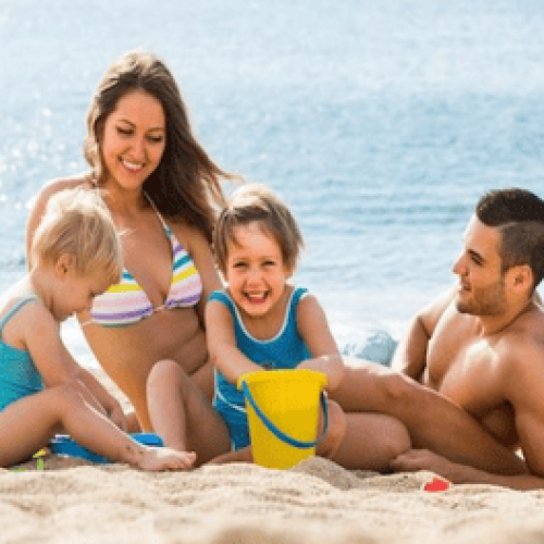 Família com duas crianças brincando alegremente na areia da praia, representando momentos de diversão típicos das Melhores praias para crianças no Espírito Santo; os pais relaxam ao lado enquanto os pequenos usam baldinhos para cavar, com o mar calmo e o clima ensolarado ao fundo