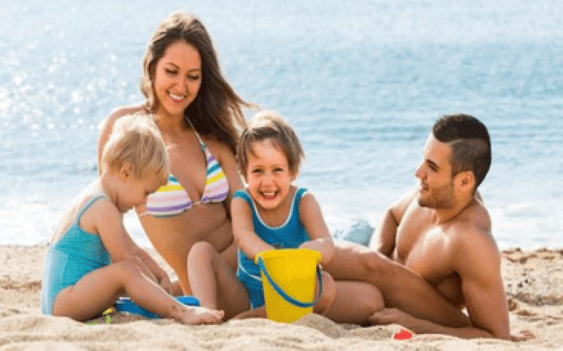 Família com duas crianças brincando alegremente na areia da praia, representando momentos de diversão típicos das Melhores praias para crianças no Espírito Santo; os pais relaxam ao lado enquanto os pequenos usam baldinhos para cavar, com o mar calmo e o clima ensolarado ao fundo