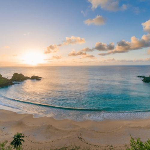 Vista panorâmica da Baía do Sancho em Fernando de Noronha, com mar azul cristalino, falésias cobertas por vegetação e o sol se pondo no horizonte, iluminando a praia de forma suave, umas das praias mais bonitas do brasil