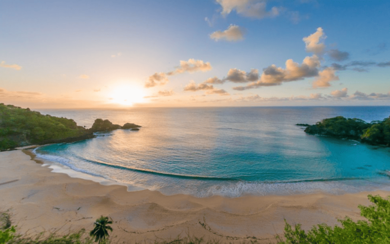 Vista panorâmica da Baía do Sancho em Fernando de Noronha, com mar azul cristalino, falésias cobertas por vegetação e o sol se pondo no horizonte, iluminando a praia de forma suave, umas das praias mais bonitas do brasil